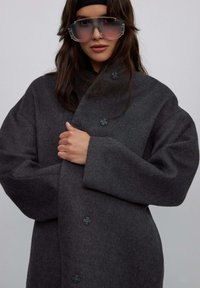 Šedý oversize vlněný kabát s velkými knoflíky, širokými rukávy a měkkou strukturou. Modelka má na sobě růžové oversize sluneční brýle.