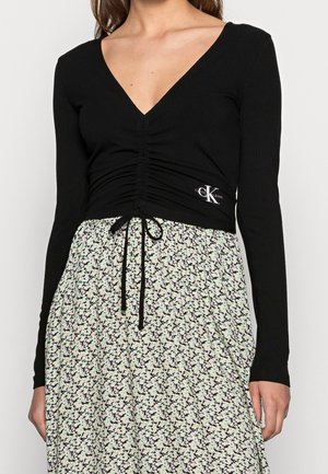 Schwarz, langärmliges Oberteil aus geripptem Stoff mit tiefem V-Ausschnitt, Raffung und Logostickerei. Kombiniert mit einem hellgrünen Rock mit floralem Muster.