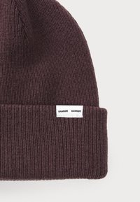 Sötétlila kötött sapka bordázott mintával. A hajtott szegélyen fehér címke hímzett „SAMSØE SAMSØE” felirattal.