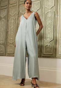Mintgrøn, ærmeløs jumpsuit med dyb V-hals og brede ben, lavet af glat stof, med en frontfald for ekstra detalje.