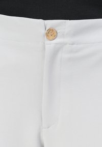 Pantalons en tissu blanc au toucher lisse, avec un bouton doré visible à la taille et un détail de poche à l'avant.