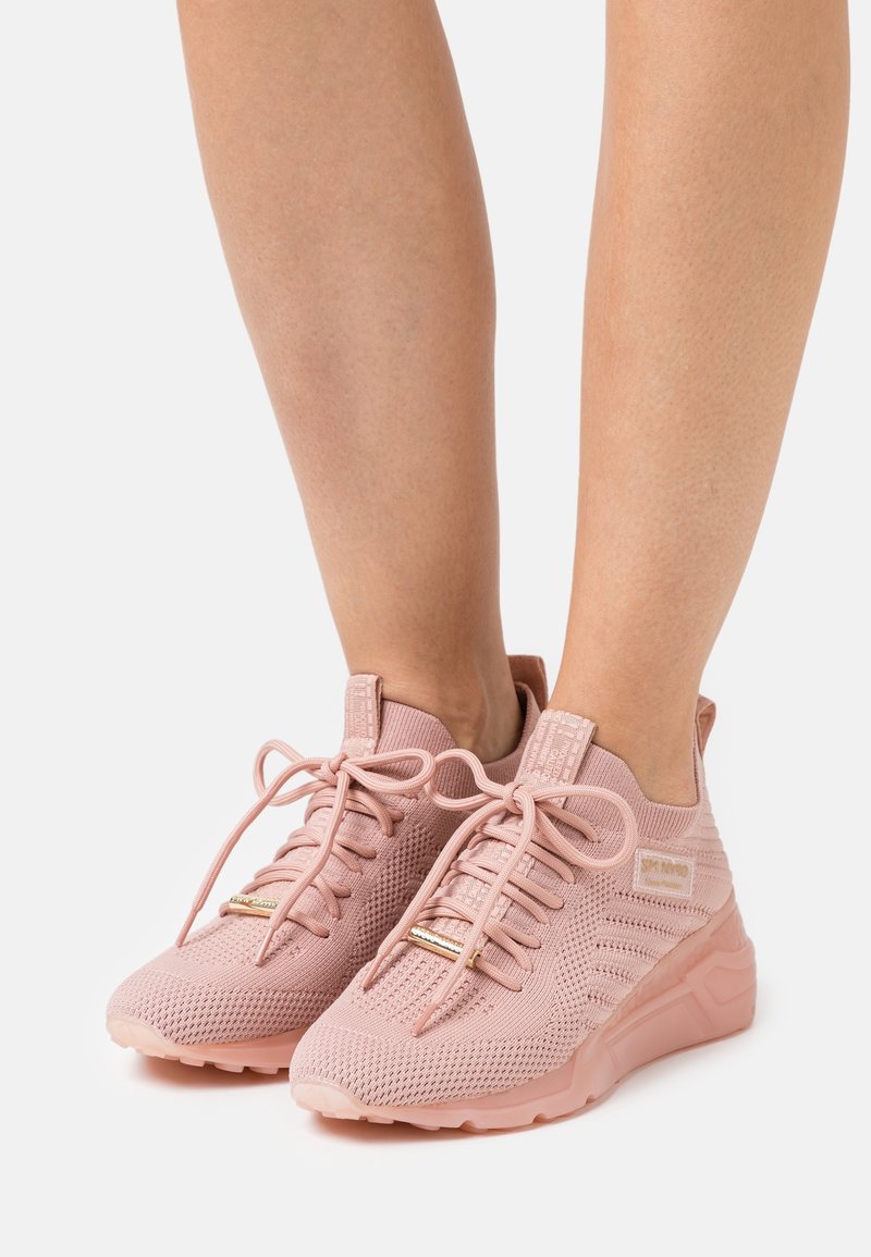 Personne portant des baskets en tricot rose avec des lacets, debout sur une surface blanche, montrant les jambes inférieures et les pieds.