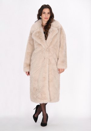 Cappotto lungo in pelliccia sintetica beige, con un ampio colletto, maniche lunghe e una texture morbida. Indossato sopra un top scuro e abbinato a tacchi neri.