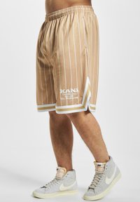 Shorts de basketball beiges avec des rayures verticales blanches, dotés d'un ourlet blanc et d'un détail de logo sur le côté gauche. Associés à des baskets gris clair.