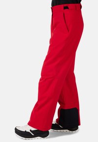 Pantaloni impermeabili rossi, vestibilità aderente, con tasche laterali e vita regolabile. Dettagli neri nella parte inferiore. Tessuto liscio e resistente.