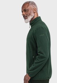 Fleecejacke in Dunkelgrün mit hohem Kragen, Reißverschluss-Taschen und gesticktem Logo-Detail am Ärmel. Glatte, weiche Textur.