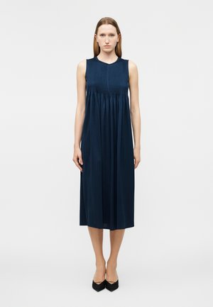 Γυναίκα που στέκεται φορώντας ένα μπλε ναυτικό αμάνικο μίντι φόρεμα με πλισέ μπούστο και μαύρα τακούνια με μυτερή μύτη, μπροστά σε μια απλή λευκή επιφάνεια.