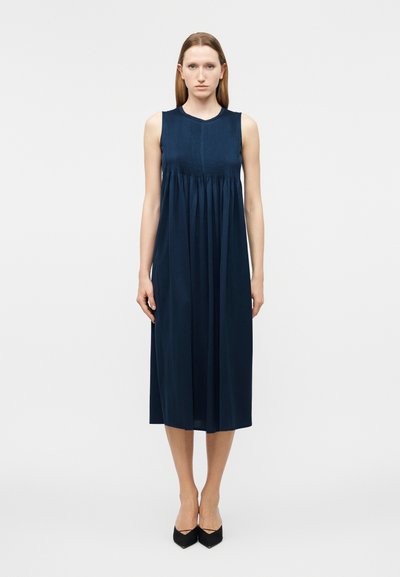 Femme debout portant une robe midi bleu marine sans manches avec un corsage plissé et des escarpins noirs à bout pointu, sur fond blanc uni.