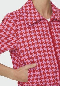 Rood en roze houndstooth patroon zip-up jack met een puntige kraag en zijzak, met een textuurstof en ribbelaccenten.