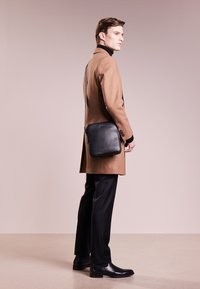 Borsa a messenger in pelle nera con chiusura a zip, forma quadrata, design sottile e branding discreto. Texture liscia e accenti hardware minimali.