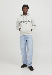 Sudadera con capucha color crema con "Originals" impreso en negro. Cuenta con un bolsillo frontal y puños acanalados, combinada con unos jeans rectos de color azul claro.