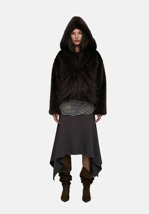 Femme portant une veste à capuche en fourrure foncée sur un haut rayé, une jupe asymétrique foncée et des bottes marron détendues, debout devant un fond uni.