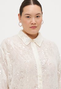 White sequined shirt with a collared neckline, sheer texture, and decorative patterns. Features a button front and oversized sleeves.