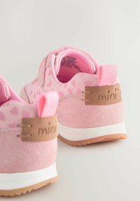Chaussures pour enfants rose avec un talon en daim texturé, motif en forme de cœur sur le tissu, une languette de traction et un détail de marque brun sur le côté.