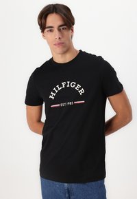 T-shirt en coton noir avec un grand logo blanc "HILFIGER" et le texte "EST. 1985" en dessous, accentué par des rayures rouges et blanches dans le design.