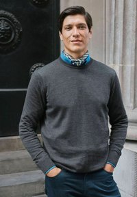 Grå tröja med långa ärmar och rund hals. En blå skjorta med krage och en mönstrad scarf syns. Slim-fit mörka byxor kompletterar outfiten.