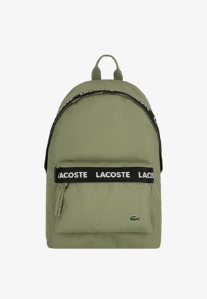 Green fabric backpack with black branded strap, zippered front pocket, and embroidered logo. Smooth texture and rounded shape.