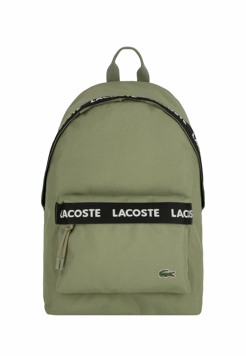 Mochila de tela verde con correa negra de marca, bolsillo frontal con cremallera y logo bordado. Textura suave y forma redondeada.