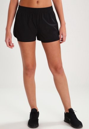 Vrouw draagt zwarte sportshorts en zwarte hardloopschoenen, staat met licht uit elkaar gespreide benen tegen een effen witte achtergrond.