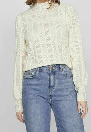 Kvinde iført en cremefarvet blondebluse med lange ærmer og bladmønstre samt højtaljede blå jeans, stående foran en ensfarvet baggrund.