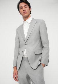 Fato cinza claro feito de tecido texturizado, apresentando um fecho de dois botões, lapelas em bico e um único bolso no peito. Acompanhado por uma camisa branca.