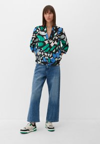 Blouse noire avec des motifs abstraits verts et bleus, décolleté en V et manches 3/4, associée à un jean large bleu clair et des baskets multicolores.