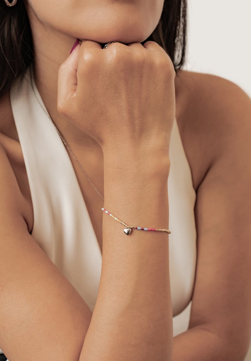 Mujer descansando la barbilla sobre el puño, lleva una pulsera de cuentas con un pequeño colgante de corazón de plata en la muñeca y una blusa blanca sin mangas.