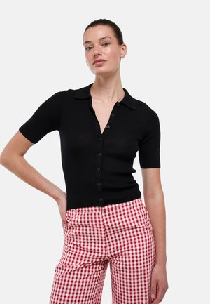 Jeune femme aux cheveux attachés, portant un haut noir à manches courtes boutonné et un pantalon à carreaux rouges et blancs, debout, main sur la hanche.