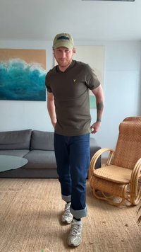 Olivgrön t-shirt med korta ärmar och en liten logotyp, tillsammans med mörk denimjeans med uppvikta muddar och vita sneakers med detaljer.