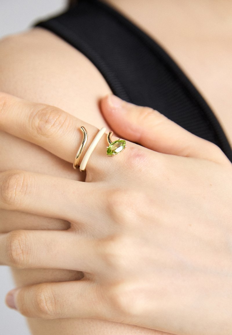 Dva prstene na ruke: jeden elegantný zlatý prsteň s vlnovým tvarom a hladký krémový prsteň, ktorý je zvýraznený zeleným drahokamom.
