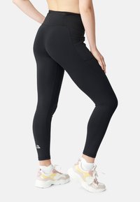 Leggings atletici neri a vita alta con una texture liscia e un design aderente. Dettaglio logo riflettente vicino alla caviglia. Indossati con sneakers pastello.