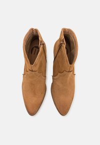 Botines de ante marrón con punta puntiaguda, bordes ondulados y cremalleras laterales, que cuentan con una suela interior texturizada.