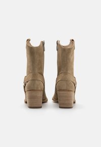 Bottines en daim tan avec une fermeture éclair sur le côté, présentant un talon empilé et un détail de sangle ajustable. Texture lisse avec un bout arrondi.