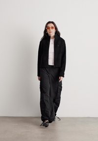 Svart teksturert jakke, rosa skjorte og svarte cargo-bukser. Tilbehør inkluderer oransje solbriller og svart fottøy. Enkel, moderne silhuett.