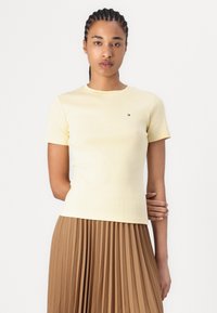 Short-sleeve yellow t-shirt made from soft fabric, featuring a small logo on the chest, paired with a pleated brown skirt.