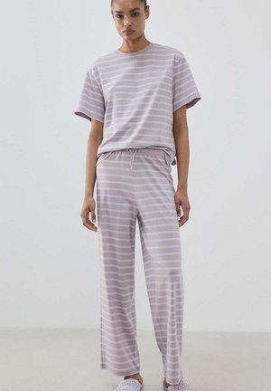 Vrouw draagt een bijpassend lichtpaars en wit gestreept kort mouwen top en broek met wijde pijpen met witte instapschoenen.