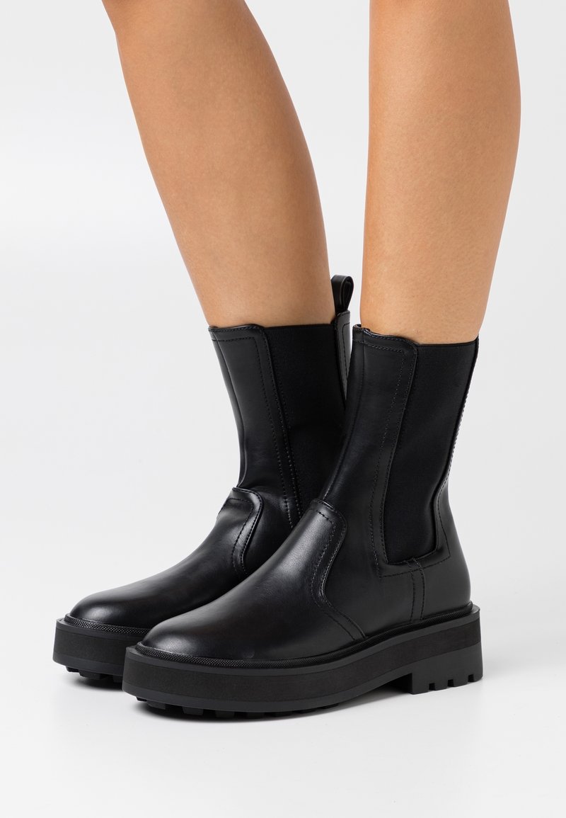 Musta nilkkabootsit, jotka on valmistettu sileästä nahasta, joustavilla sivupaneeleilla, pyöristetyllä varvashuolella, paksulla kumipohjalla ja hienovaraisilla ompeluyksityiskohdilla.