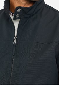 Men's black jacket featuring a high collar, metal snap buttons, a front zip closure, and a textured fabric.