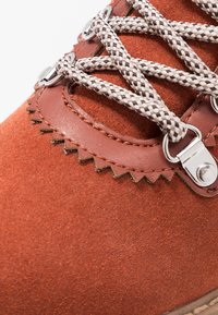 Zapato de ante color óxido con una superficie texturizada, que presenta ojales metálicos y cordones trenzados. El ribete de piel tiene un detalle de borde en zigzag.