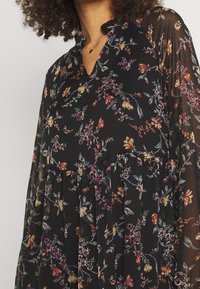 Zwarte bloemen jurk met lichte, doorschijnende lange mouwen, een gathered taille en een V-hals met een kleine kraag en kleurrijke bloemenpatronen.