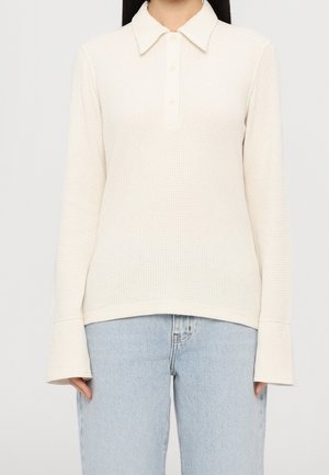 Femme portant un polo à manches longues en maille côtelée crème avec des poignets prolongés et un jean taille haute bleu clair.