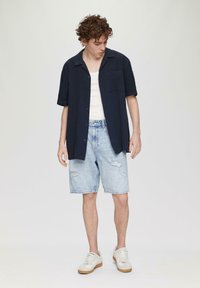 Marineblauwe korte mouwen overhemd over een wit tanktop, gedragen met lichtblauwe versleten denimshorts en witte sneakers met tan kleurige zolen.