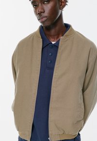 Giacca bomber marrone in tessuto, con zip frontale, polsini a coste e colletto foderato. Indossata sopra una polo blu navy con bottoni.