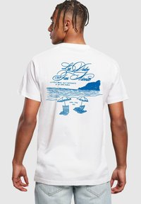 White cotton t-shirt featuring a blue graphic print on the back with beach chairs, umbrellas, and the text “La Dolce Far Niente.”