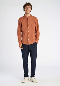 Roestkleurig overhemd met kraag en borstzak, gecombineerd met donkerblauwe broek en witte sneakers. Glad stofmateriaal.