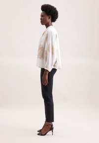 Blusa bianca a maniche lunghe con motivi floreali dorati, abbinata a jeans skinny neri e sandali neri con tacco. Tessuto morbido, vestibilità rilassata.