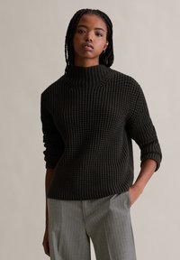 Dunkelgrüner Strickpullover mit hohem Kragen, strukturiertem Design und Dreiviertelärmeln, kombiniert mit hellgrauen, gestreiften Hosen.