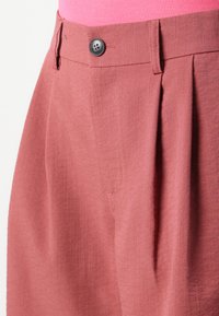 Pantalon plissé rose en tissu léger. Doté d'une fermeture à bouton noir et d'une texture lisse. Taille cintrée avec poches latérales.