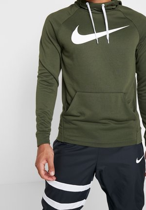 Personne portant un sweat à capuche Nike vert olive avec un logo blanc et un pantalon de sport noir avec des rayures horizontales blanches et le logo Nike sur la cuisse gauche.