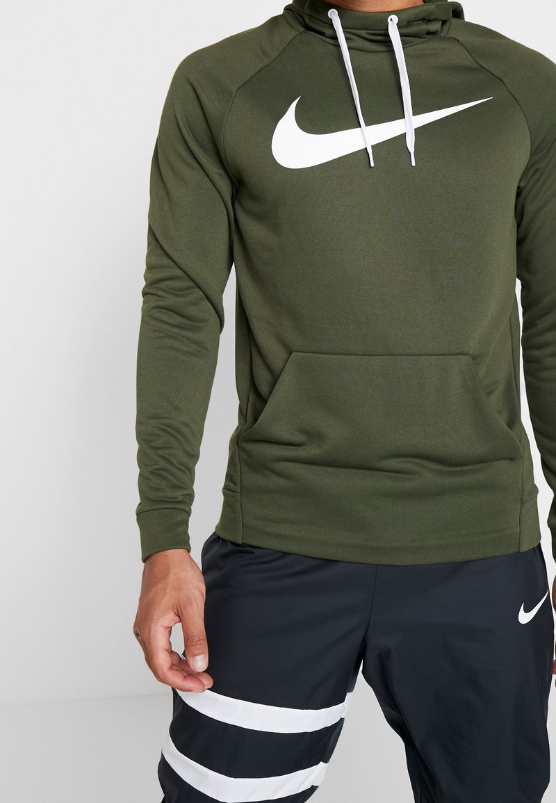 Personne portant un sweat à capuche Nike vert olive avec un logo blanc et un pantalon de sport noir avec des rayures horizontales blanches et le logo Nike sur la cuisse gauche.